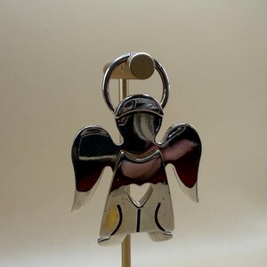 May include: A shiny silver angel-shaped keychain. The angel has a halo, wings, and a heart-shaped cutout. The keychain is attached to a silver ring. The angel is displayed on a wooden stand.