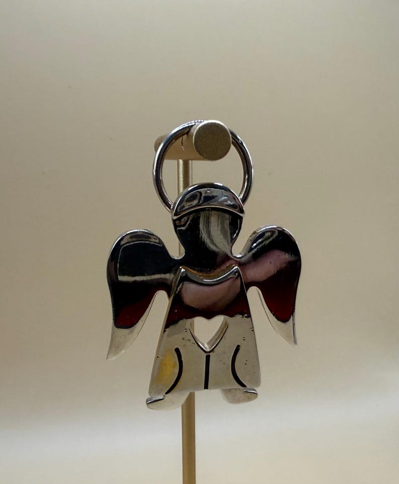 May include: A shiny silver angel-shaped keychain. The angel has a halo, wings, and a heart-shaped cutout. The keychain is attached to a silver ring. The angel is displayed on a wooden stand.