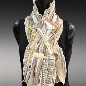May include: A distinctive, handmade scarf with a layered design. It features various textures and colours, including cream, beige, and muted pink. The scarf is adorned with gold-toned metal accents and a leather patch, creating a bohemian aesthetic.