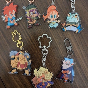 Puede incluir: Ocho llaveros de personajes de dibujos animados coloridos con cadenas plateadas y doradas. Los personajes incluyen un guerrero pelirrojo, una rana, un robot, una mujer rubia, un vampiro, una mujer con una corona, una mujer con un turbante y un hombre con un sombrero.