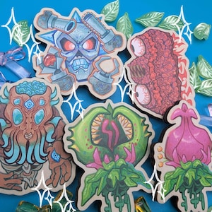 Op de afbeelding: Een verzameling kleurrijke, cartoonachtige houten stickers met verschillende monster- en wezendesigns. De stickers bevatten een schedel met een kettingzaag, een wezen met meerdere ogen en andere unieke karakters. De achtergrond is helderblauw met decoratieve accenten.