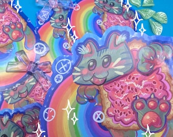 Puede incluir: Una colección de pegatinas coloridas con un diseño de gato de dibujos animados. El gato tiene pelaje gris y rosa, un fondo de arcoíris y una pata rosa. Las pegatinas tienen una estética caprichosa y juguetona, con un fondo azul.
