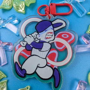 以下が含まれることがあります： 青と白のユニフォームを着てバットを振る、漫画のバッターをフィーチャーした野球をテーマにしたキーホルダー。 キーホルダーには赤い留め金があり、装飾用のプラスチック製の葉とリボンに囲まれています。