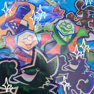 Puede incluir: Una colección de pegatinas coloridas de estilo caricatura con varios diseños, incluyendo una cara guiñando un ojo, una rana con traje y un lazo. Las pegatinas tienen un acabado brillante y están dispuestas sobre un fondo azul claro.