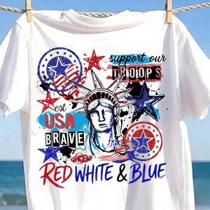 Könnte beinhalten: Ein weißes T-Shirt mit einem Grafikdruck der Freiheitsstatue, umgeben von roten, weißen und blauen Sternen und Kreisen. Der Text lautet support our TROOPS, est USA BRAVE und RED WHITE & BLUE in einer Mischung aus verschiedenen Schriftarten.