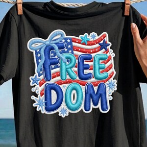 Könnte beinhalten: Ein schwarzes T-Shirt mit einem Grafikdesign einer stilisierten amerikanischen Flagge und einer hellblauen Schleife oben links. Das Wort FREEDOM ist in großen, aufgeblähten, dunkel- und hellblauen Buchstaben gedruckt, umgeben von kleinen blauen Sternen.