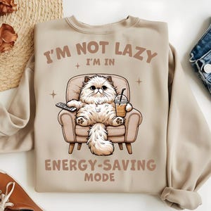 Op de afbeelding: Beige sweatshirt met de tekst "I'M NOT LAZY I'M IN ENERGY-SAVING MODE". De afbeelding toont een pluizige witte kat die in een stoel zit, met een afstandsbediening en een drankje. De kat is de centrale focus van het ontwerp.