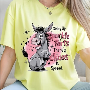 Pode incluir: Camiseta amarelo claro com um desenho de burro de desenho animado e o texto "Giddy Up Sparkle Farts There's Chaos to Spread". O burro &eacute; cinza com crina e cauda pretas, em um fundo rosa.