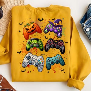 May include: Mustard yellow sweatshirt featuring Halloween-themed video game controller designs. The controllers are decorated with images of a jack-o'-lantern, witch hat, Frankenstein, and other spooky elements. Bats and spiderwebs are also included in the design.