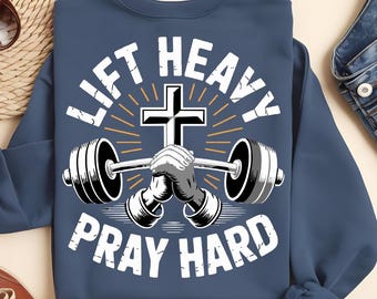 May include: Navy blue sweatshirt with the text "LIFT HEAVY PRAY HARD". The design features a barbell with hands clasped in prayer, a cross, and radiating lines. The sweatshirt is a comfortable, casual style.