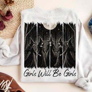 May include: White sweatshirt with a black and white illustration of five witches in a forest under a full moon. The text "Girls Will Be Girls" is printed below the image. The design has a spooky, gothic theme.