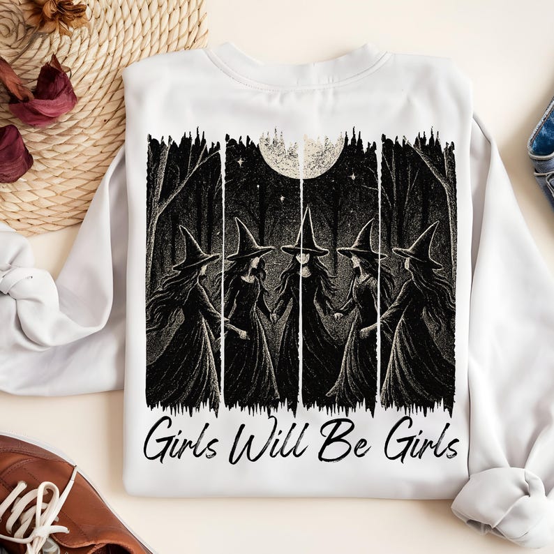 May include: White sweatshirt with a black and white illustration of five witches in a forest under a full moon. The text "Girls Will Be Girls" is printed below the image. The design has a spooky, gothic theme.