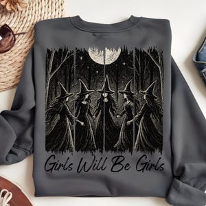 May include: Dark grey sweatshirt with a black and white illustration of five witches in a forest under a full moon. The text "Girls Will Be Girls" is printed below the image. A comfortable, long-sleeved top.