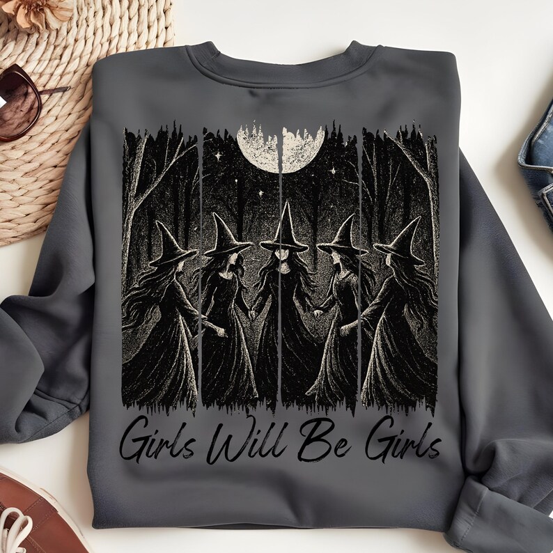 May include: Dark grey sweatshirt with a black and white illustration of five witches in a forest under a full moon. The text "Girls Will Be Girls" is printed below the image. A comfortable, long-sleeved top.