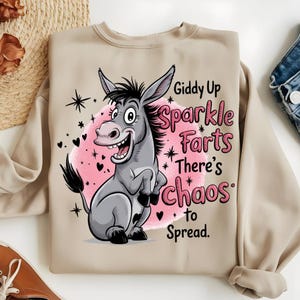 Pode incluir: Camisola bege com um desenho de burro de desenho animado e o texto "Giddy Up Sparkle Farts There's Chaos to Spread." O burro &eacute; cinzento com um c&iacute;rculo rosa, estrelas e cora&ccedil;&otilde;es pretos.