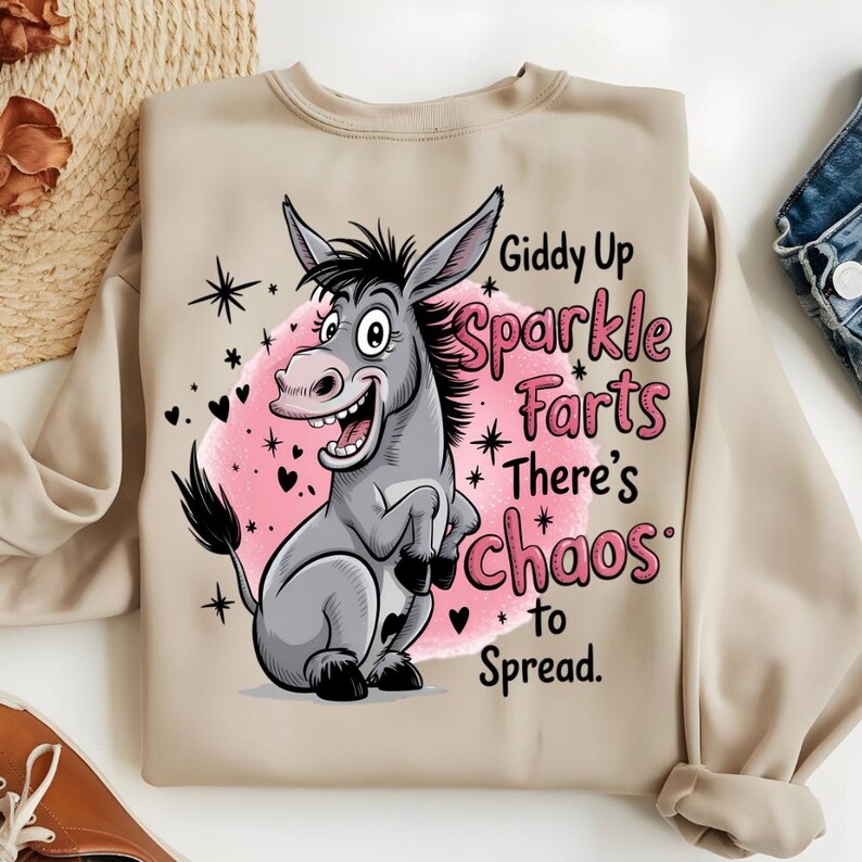 Pode incluir: Camisola bege com um desenho de burro de desenho animado e o texto "Giddy Up Sparkle Farts There's Chaos to Spread." O burro &eacute; cinzento com um c&iacute;rculo rosa, estrelas e cora&ccedil;&otilde;es pretos.