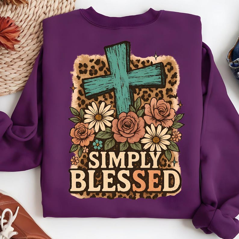 May include: A purple sweatshirt featuring a teal cross, floral arrangement, and the words "SIMPLY BLESSED." The design includes roses, daisies, and a leopard print background. The sweatshirt is a solid purple color.