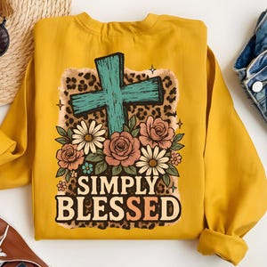 May include: Mustard yellow sweatshirt with a turquoise cross design, surrounded by flowers and a leopard print background. The text "SIMPLY BLESSED" is printed below the floral arrangement. The sweatshirt has long sleeves and a crew neck.