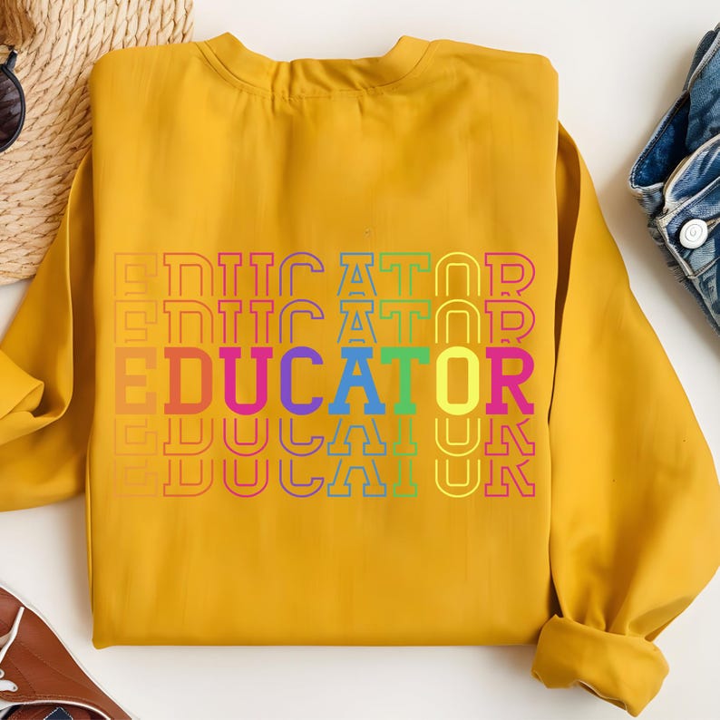 May include: Mustard yellow sweatshirt with the word "EDUCATOR" in a rainbow gradient design. The text is stacked vertically, with each word slightly offset. The sweatshirt has long sleeves and a crew neck.