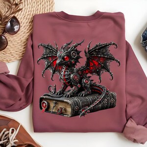 May include: A maroon sweatshirt featuring a detailed illustration of a steampunk dragon. The dragon, with red and black accents, is perched atop a book. The design incorporates gears and mechanical elements, creating a fantasy-themed aesthetic.