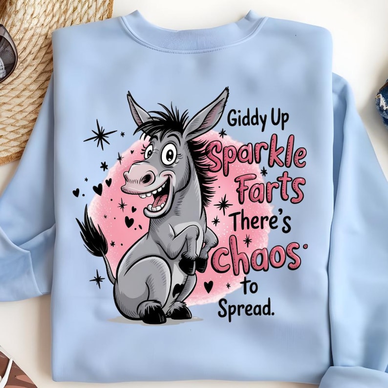 Pode incluir: Camisola azul clara com uma ilustra&ccedil;&atilde;o de um burro de desenho animado. O burro &eacute; cinzento com detalhes pretos, sobre um fundo rosa. O texto na camisa diz "Giddy Up Sparkle Farts There's Chaos to Spread."