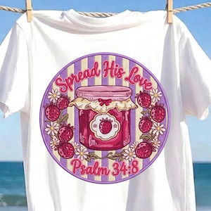 May include: A white t-shirt featuring a circular graphic with a purple and yellow striped background. The design shows a jar of strawberry jam surrounded by strawberries and daisies, with the text "Spread His Love" and "Psalm 34:8" in pink.