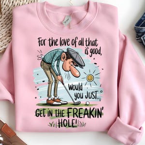 Puede incluir: Sudadera rosa claro con un dise&ntilde;o de golf. El dise&ntilde;o presenta un golfista de dibujos animados y el texto "For the love of all that is good, would you JUST, GET IN THE FREAKIN' HOLE!". Incluye una pelota de golf y un sol.