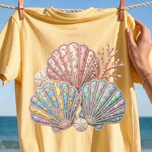 Puede incluir: Una camiseta de manga corta de color amarillo pálido con un gráfico central de tres conchas marinas grandes y una rama de coral. Las conchas tienen patrones florales y geométricos en rosa, azul, amarillo y verde, decoradas con cuentas perladas.