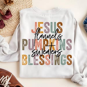 May include: White sweatshirt with the words "JESUS, flannel, PUMPKINS, sweaters, BLESSINGS" in various colors. The text is in a mix of fonts and styles, creating a visually appealing design. The sweatshirt is a casual, comfortable style.