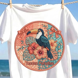 May include: A white t-shirt featuring a circular graphic with a toucan perched among hibiscus flowers and tropical leaves. The design includes two peach-colored bows on the sides and the text Toucan Paradise printed in a bold, dark font at the bottom.