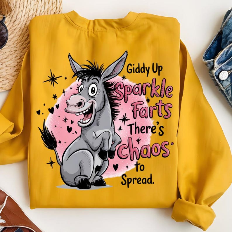 Pode incluir: Camisola mostarda com um desenho de burro de desenho animado e o texto "Giddy Up Sparkle Farts There's Chaos to Spread." O burro &eacute; cinzento com um fundo rosa e detalhes em preto. O design inclui estrelas e cora&ccedil;&otilde;es.
