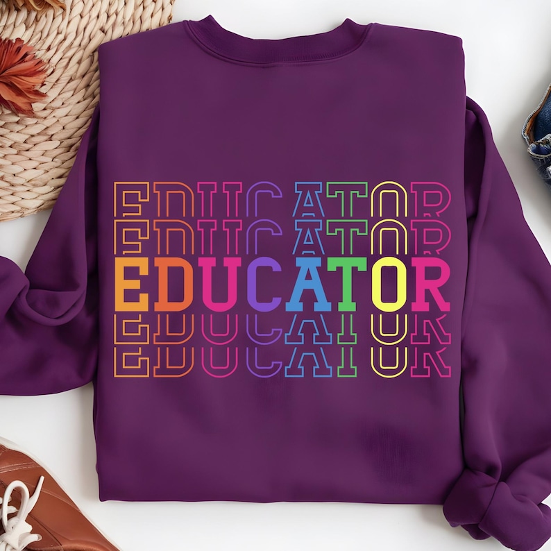 May include: A purple sweatshirt with the word "EDUCATOR" in a rainbow-colored, stacked design. The word is outlined in a contrasting color, creating a bold and eye-catching graphic. The sweatshirt is a crew neck style.