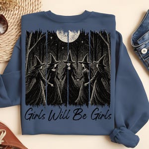 May include: A blue sweatshirt with a graphic depicting five witches in black dresses and pointed hats, holding hands in a forest under a full moon. The text "Girls Will Be Girls" is printed below the graphic.