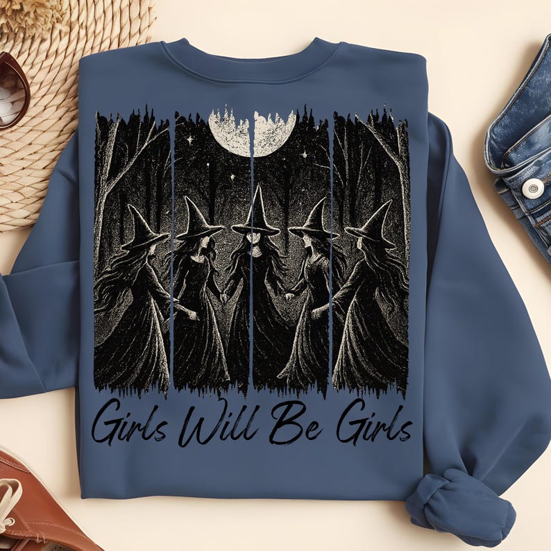 May include: A blue sweatshirt with a graphic depicting five witches in black dresses and pointed hats, holding hands in a forest under a full moon. The text "Girls Will Be Girls" is printed below the graphic.