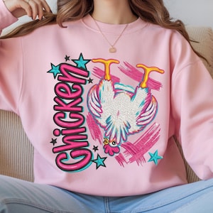 Peut inclure: Un sweat-shirt à col rond rose clair avec une impression graphique représentant un poulet blanc aux pattes jaunes, positionné à l'envers. Le motif inclut le mot "Chicken" en grandes lettres roses, agrémenté d'étoiles turquoise et de traits de pinceau roses.