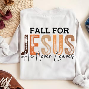 Puede incluir: Sudadera blanca con el texto "FALL FOR JESUS He Never Leaves" en letras negras, marrones y naranjas. La sudadera es de cuello redondo y manga larga.