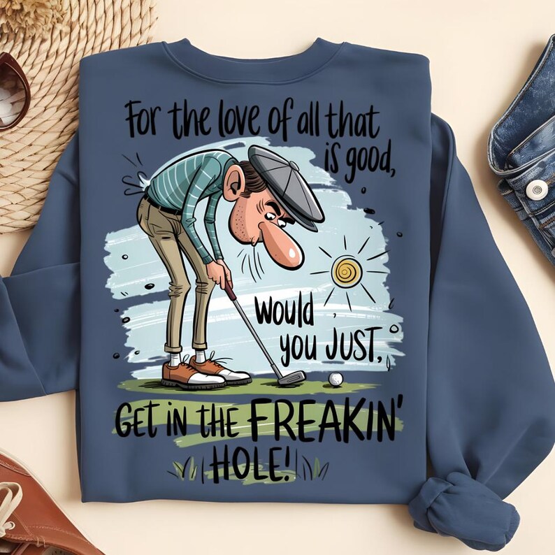 Puede incluir: Sudadera azul marino con un gr&aacute;fico humor&iacute;stico sobre el golf. La ilustraci&oacute;n muestra a un golfista con el texto: "For the love of all that is good, would you JUST, GET IN THE FREAKIN' HOLE!" Tambi&eacute;n se muestra una pelota de golf y un sol.