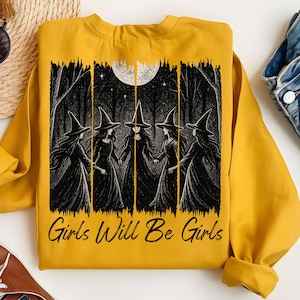 May include: Mustard yellow sweatshirt with a black and white graphic of five witches in a forest under a full moon. The text "Girls Will Be Girls" is printed below the graphic. The sweatshirt has long sleeves.