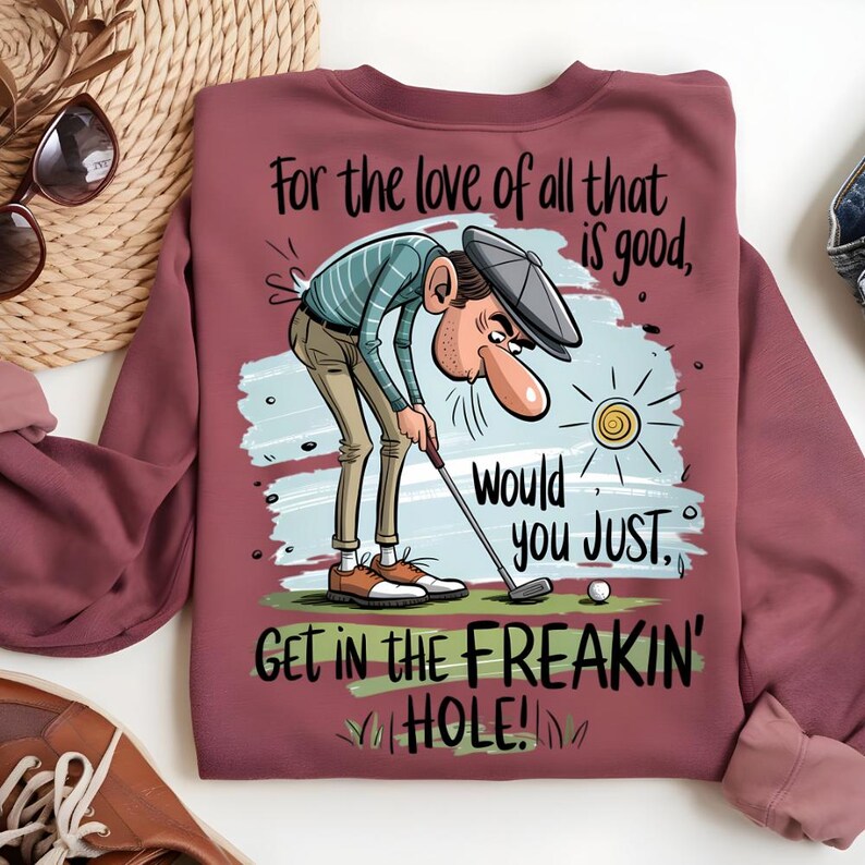 Puede incluir: Sudadera burdeos con un gr&aacute;fico tem&aacute;tico de golf. La ilustraci&oacute;n representa a un golfista con el texto "For the love of all that is good, Would you JUST GET IN THE FREAKIN' HOLE!" Una pelota de golf y un sol tambi&eacute;n est&aacute;n en el dise&ntilde;o.