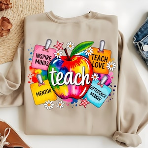 Może przedstawiać: Beżowa bluza z kolorową grafiką jabłka. Jabłko jest w stylu tie-dye z białym napisem "teach". Wokół jabłka znajdują się kolorowe etykiety z napisami "Inspire Minds", "Teach Love", "Mentor" i "Difference Maker".