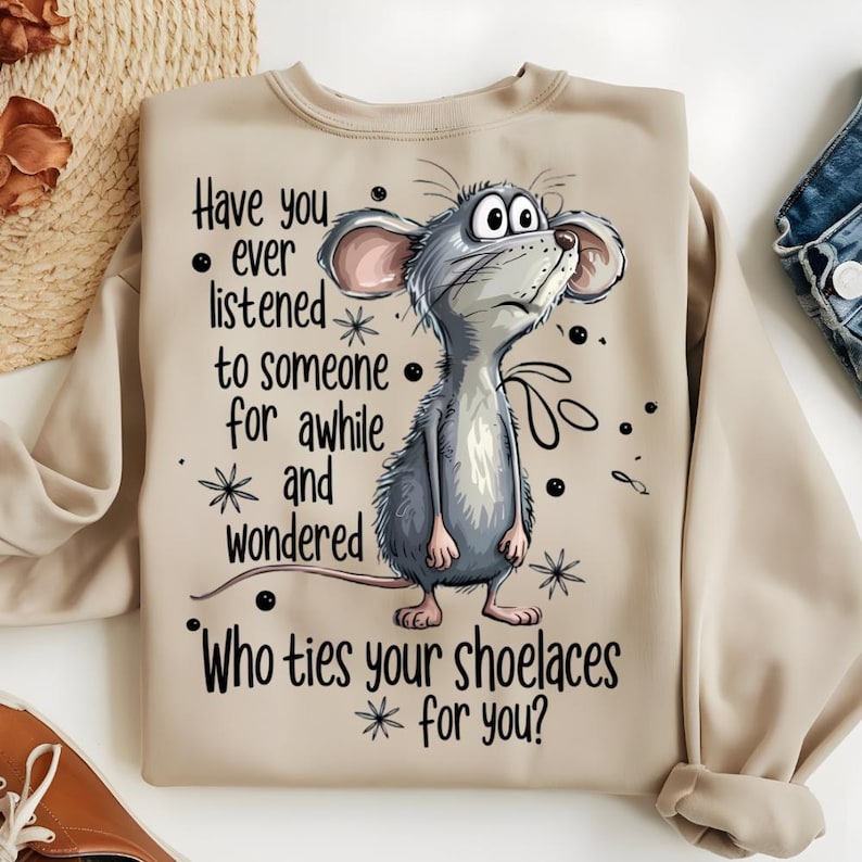 May include: Beige sweatshirt featuring a cartoon mouse with large eyes, looking upwards. The text on the shirt reads: "Have you ever listened to someone for awhile and wondered, Who ties your shoelaces for you?" The design includes stars and dots.