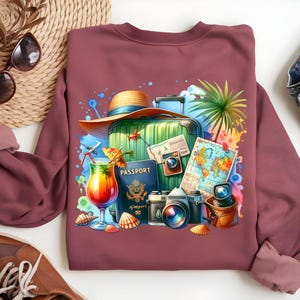 Puede incluir: Sudadera burdeos con un gr&aacute;fico de tem&aacute;tica de viajes. El dise&ntilde;o incluye una maleta verde, pasaporte, c&aacute;maras, un c&oacute;ctel, un mapa y un sombrero de paja. Conchas marinas y una hoja de palma completan el tema de las vacaciones.