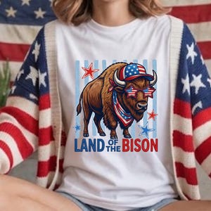 May include: A white t-shirt featuring a graphic of a brown bison wearing a baseball cap and star-shaped sunglasses with American flag patterns. The bison wears a red, white, and blue bandana. The text LAND OF THE BISON is printed below in blue and red.