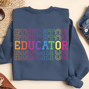 May include: A navy blue sweatshirt with the word "EDUCATOR" printed in a repeating, colorful design. The word is stacked vertically, with each letter outlined in a different color, including orange, pink, and yellow.