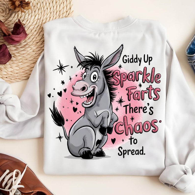 Pode incluir: Camisola branca de manga comprida com um desenho de burro de desenho animado. O burro &eacute; cinzento com detalhes pretos, sobre um fundo rosa. O texto diz "Giddy Up Sparkle Farts There's Chaos to Spread."