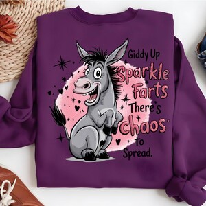 Pode incluir: Um moletom roxo com um desenho de um burro de desenho animado. O burro &eacute; cinza com uma crina preta e est&aacute; sorrindo. O texto no moletom diz "Giddy Up Sparkle Farts There's Chaos to Spread". O gr&aacute;fico est&aacute; em um fundo rosa com cora&ccedil;&otilde;es e estrelas.