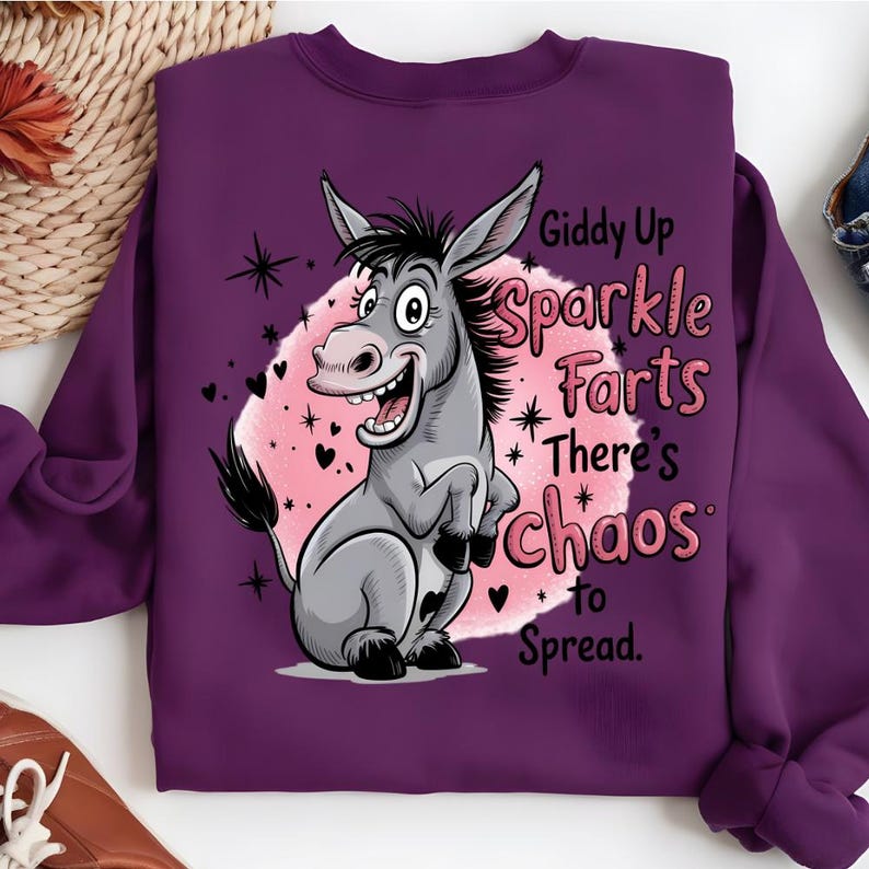 Pode incluir: Um moletom roxo com um desenho de um burro de desenho animado. O burro &eacute; cinza com uma crina preta e est&aacute; sorrindo. O texto no moletom diz "Giddy Up Sparkle Farts There's Chaos to Spread". O gr&aacute;fico est&aacute; em um fundo rosa com cora&ccedil;&otilde;es e estrelas.