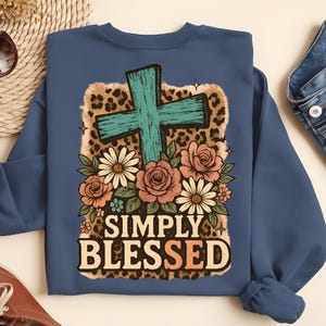 May include: A blue sweatshirt featuring a teal cross design with floral accents and the words "SIMPLY BLESSED." The design includes daisies, roses, and a leopard print background. The sweatshirt is a solid blue color.