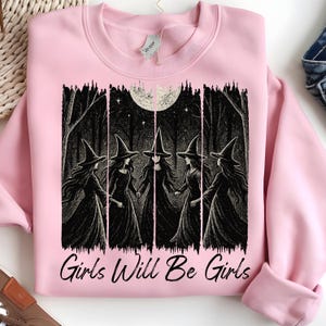 May include: A pale pink sweatshirt with a black and white illustration of five witches in a forest under a full moon. The text "Girls Will Be Girls" is printed below the graphic. The sweatshirt has a crew neck and long sleeves.
