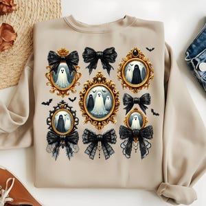 May include: Beige sweatshirt featuring framed ghost portraits and black bows. The gold frames contain white ghosts with black eyes and mouths. Black bats are scattered around the frames. The bows are black and some have lace detailing.